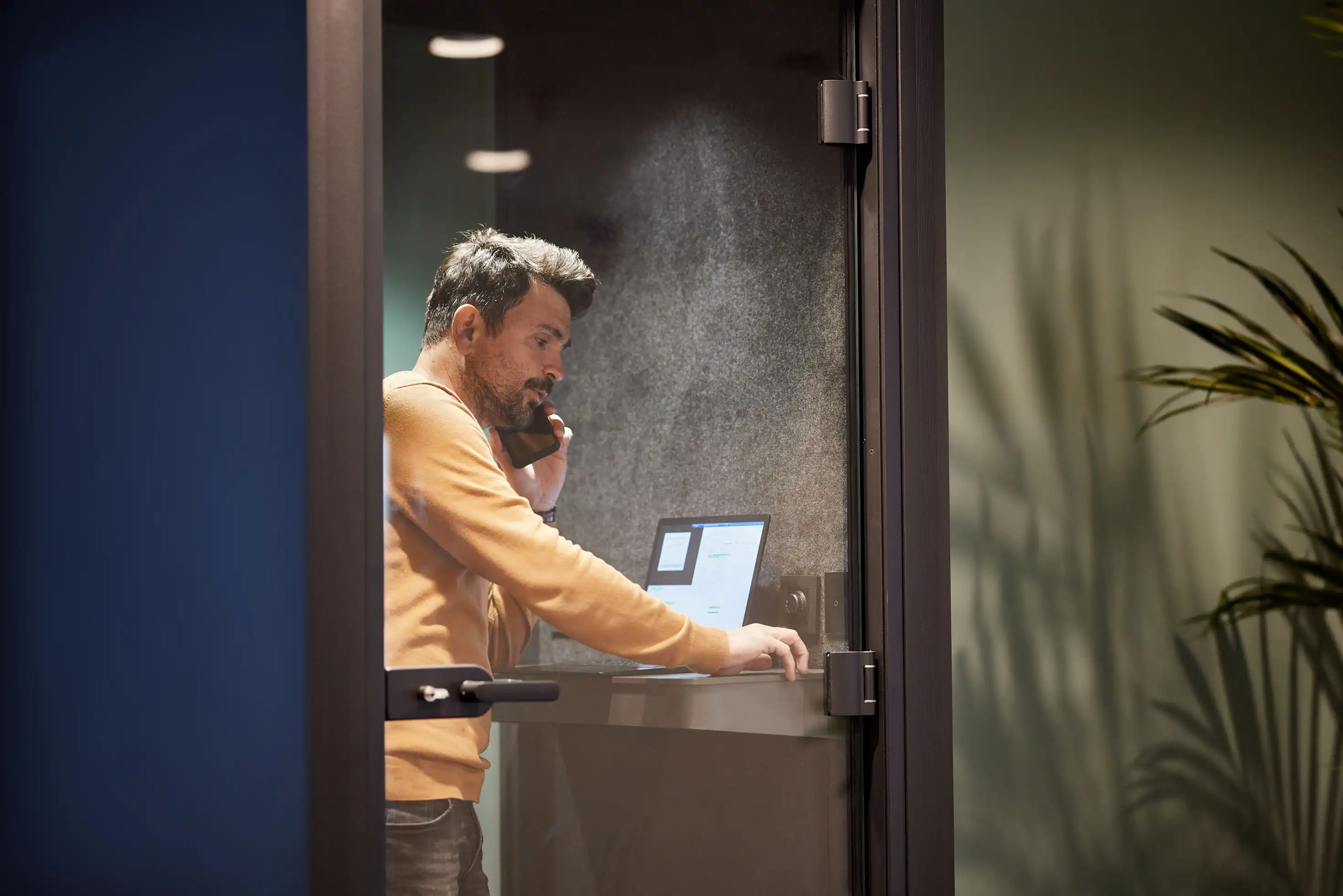 En man står inne i ett privat kontorsbås och pratar i telefon och arbetar på en bärbar dator. Rummet har glasväggar.