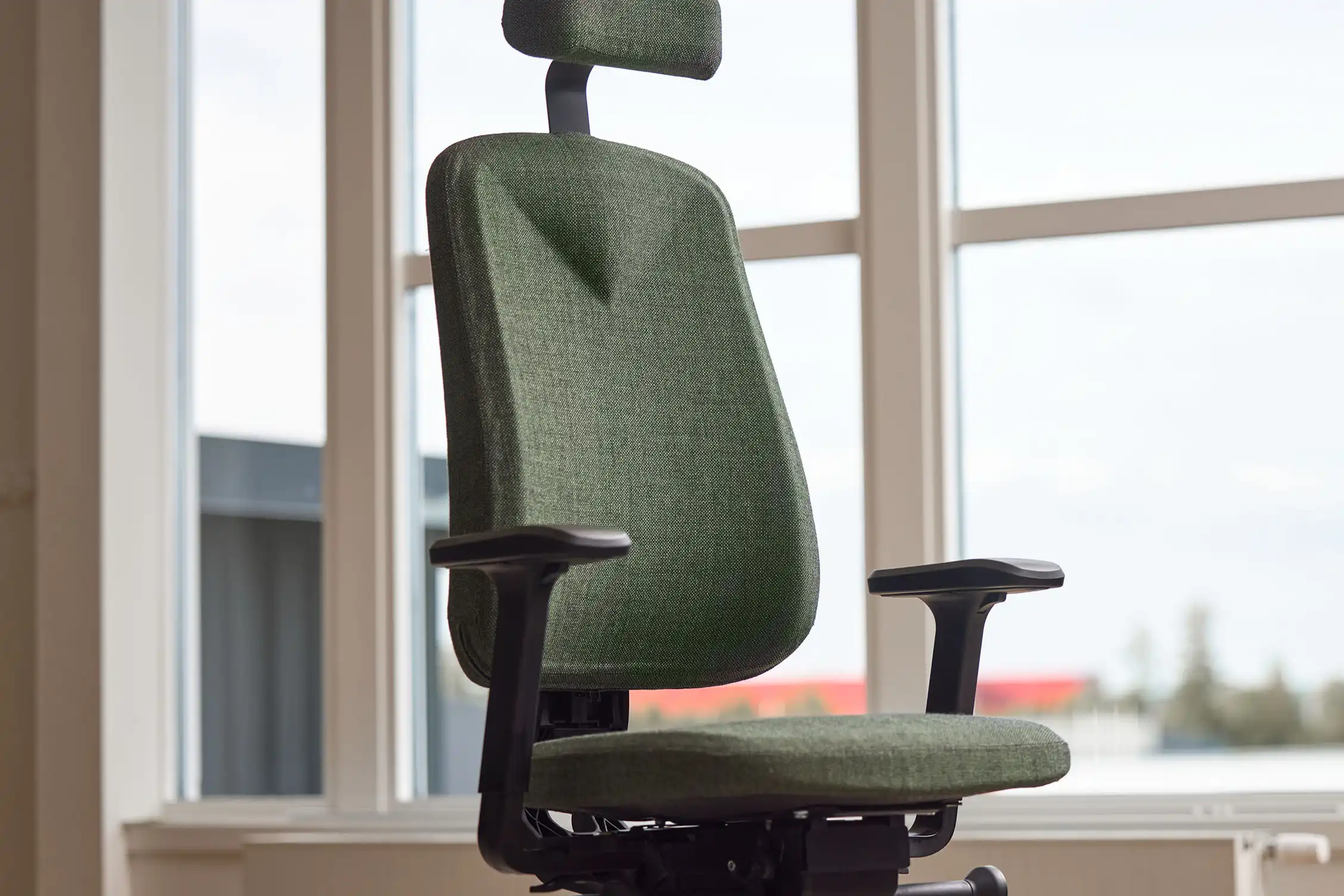 En ergonomisk kontorsstol med grönt tyg och nackstöd är placerad nära stora fönster med gott om naturligt ljus. Stolen har justerbara armstöd.