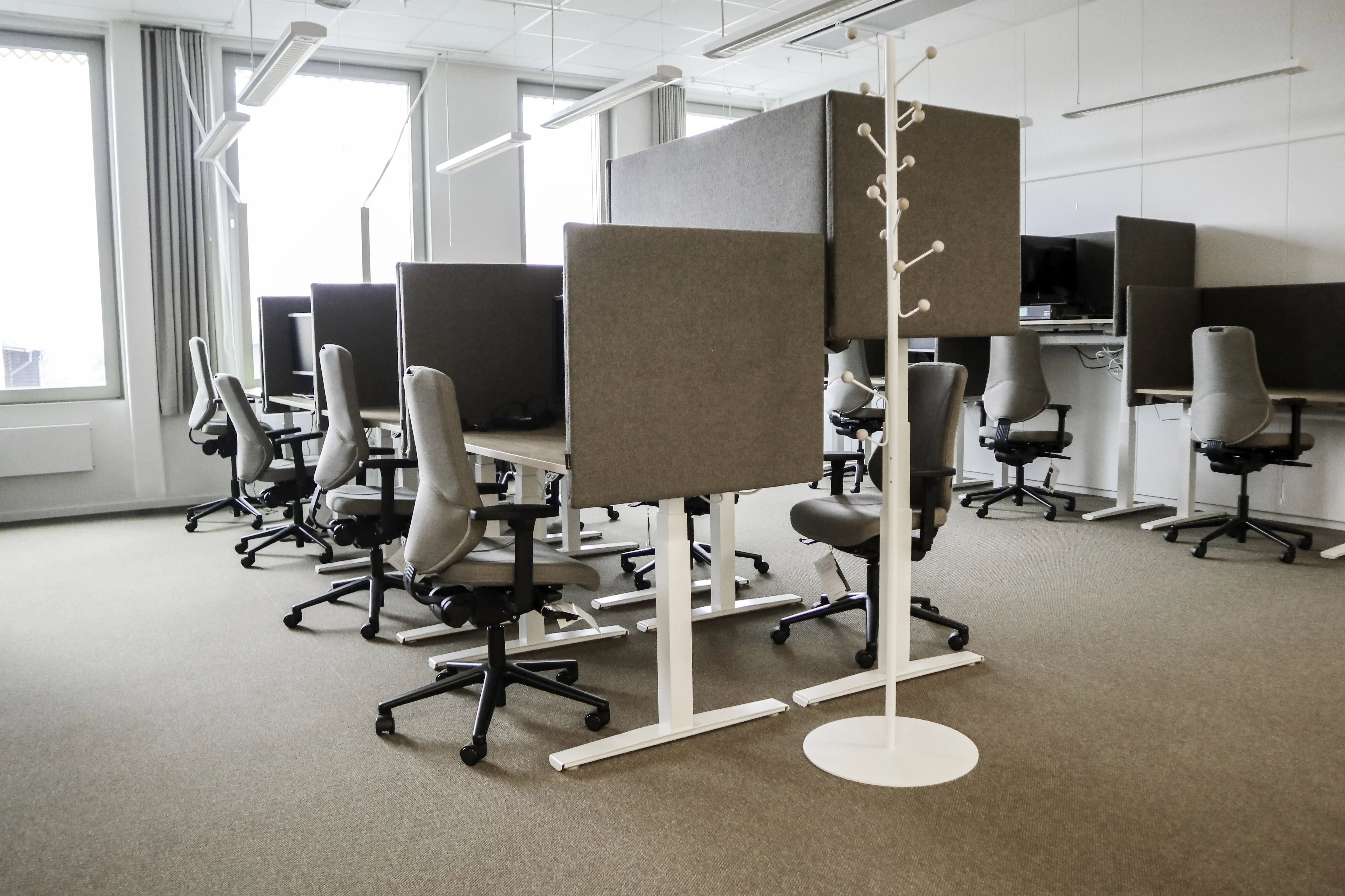 En kontorslokal med avdelade skrivbord och ergonomiska kontorsstolar. Skiljeväggarna är klädda i grått tyg. Stora fönster till vänster ger naturligt ljus i rummet.